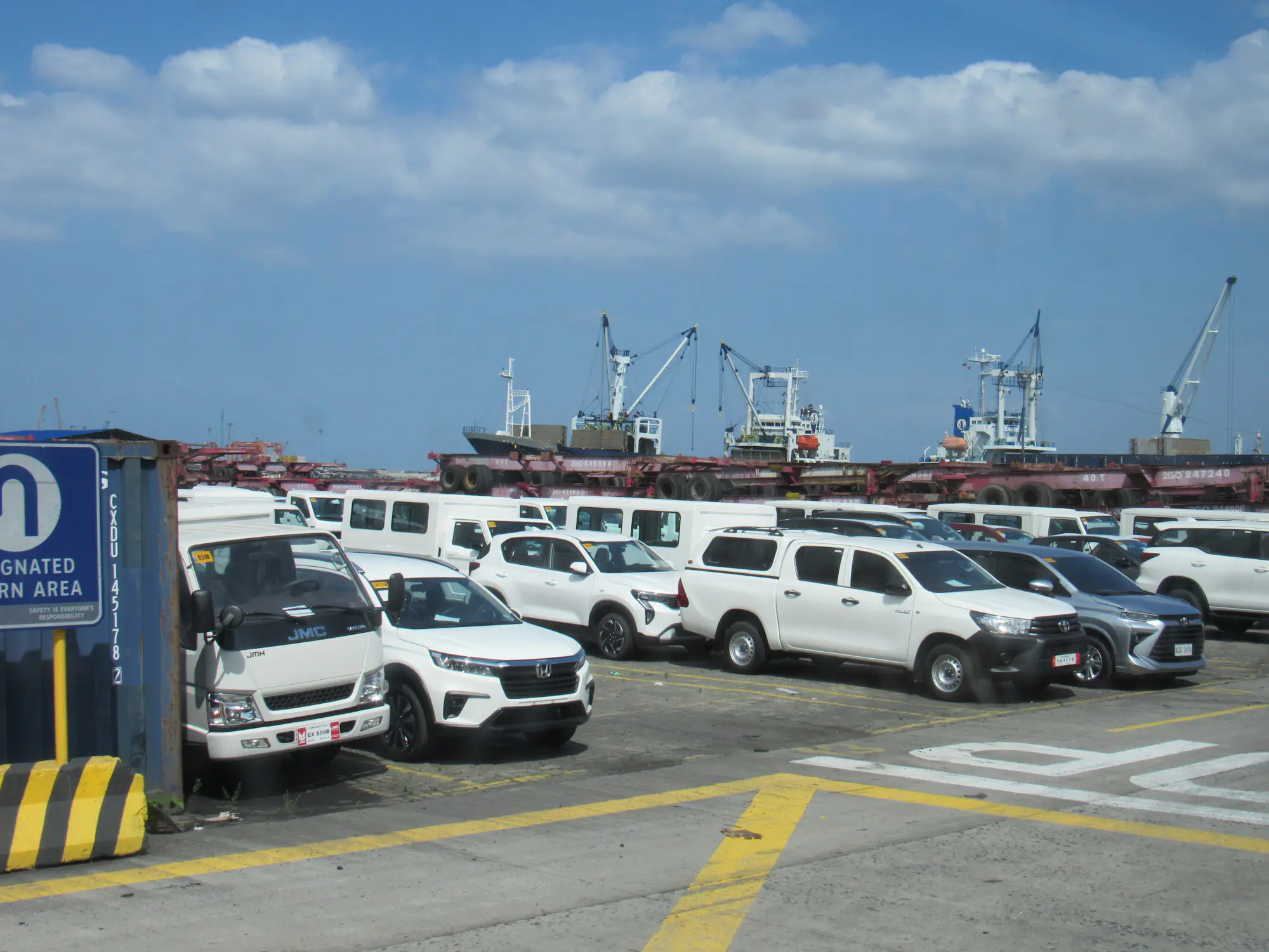 Filas de vehículos nuevos en un puerto, con las grúas de los barcos al fondo